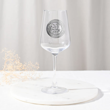 Clear stemmed wine glass with a round silver "90th Birthday" badge on the bowl, tall slim stem, shown on a marble coaster with a neutral background.