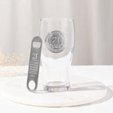 Clear pint beer glass with raised silver "21st Birthday" badge and star detail, paired with a matching stainless bottle opener engraved with milestone stats, set on a marble tray.