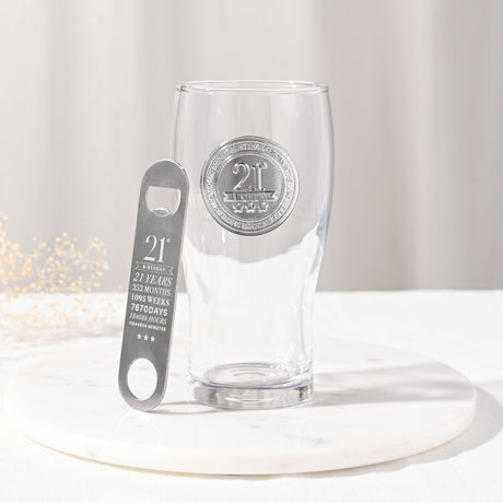 Clear pint beer glass with raised silver "21st Birthday" badge and star detail, paired with a matching stainless bottle opener engraved with milestone stats, set on a marble tray.