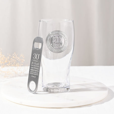 Clear tulip-shaped pint glass with silver "30th Birthday" badge and stars, beside a matching silver bottle opener engraved with 30th birthday milestones, on a light background.