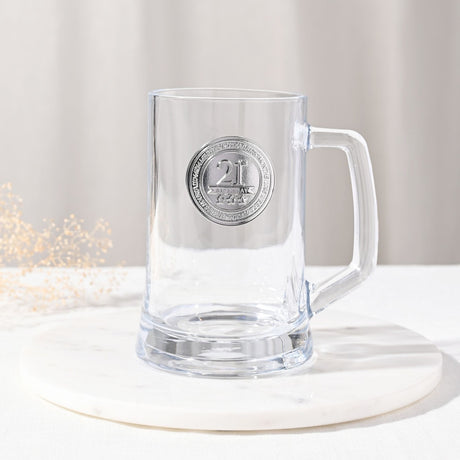 Clear glass tankard with a round silver "21" birthday badge on the front and a thick, squared handle, set on a marble coaster against a light background.