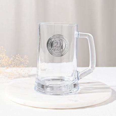 Clear glass tankard beer mug, 650ml, with a round silver-coloured 30th birthday badge, straight sides, thick base and squared handle, shown on a marble coaster against a neutral background.