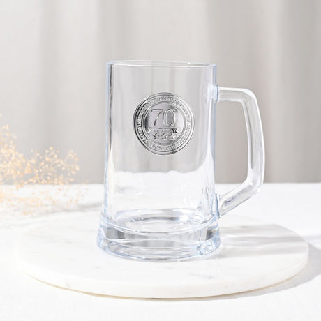 Clear glass tankard beer mug with a sturdy handle, featuring a round silver-coloured badge reading "70th Birthday" with stars, set on a marble surface against a neutral background.
