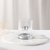 Round clear glass whisky tumbler with a raised silver 3D "30" and "THIRTY" lettering on its silver base, displayed on a marble tray with a soft neutral background.