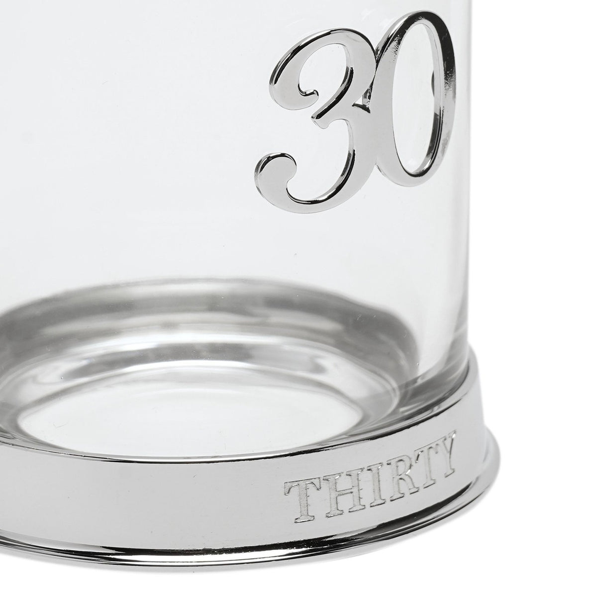 Close-up of a round clear glass whisky tumbler with a raised 3D silver "30" on the side and "THIRTY" engraved around its silver base, on a white background.