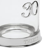 Close-up of a round clear glass whisky tumbler with a raised 3D silver "30" on the side and "THIRTY" engraved around its silver base, on a white background.