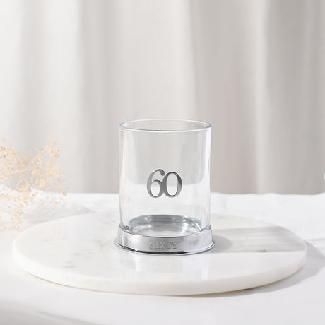 Clear glass whisky tumbler with raised silver 3D "60" numeral and a silver base engraved "SIXTY", displayed on a round marble tray against a light curtain backdrop.