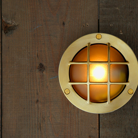 Muara brass outdoor bulkhead light, 14cm round fitting with grid grille over frosted glass dome, screw-fixed ring, mounted on dark wood panelling, lit bulb visible.