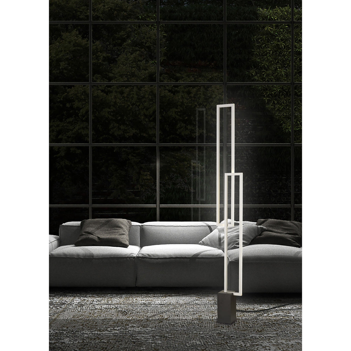 A modern living room with a light gray sofa, dark cushions, and the Mural LED Floor Lamp 48W 3000K in matt white stands by large glass windows overlooking lush greenery. A textured, patterned rug covers the floor.