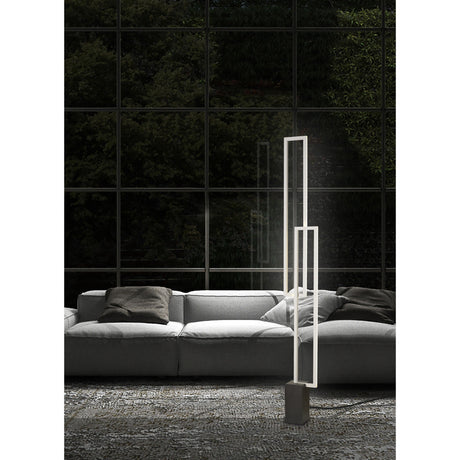 A modern living room with a light gray sofa, dark cushions, and the Mural LED Floor Lamp 48W 3000K in matt white stands by large glass windows overlooking lush greenery. A textured, patterned rug covers the floor.
