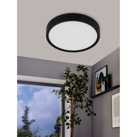 Musurita LED flush ceiling light, 34cm round, black surround with white plastic diffuser, shown mounted on a light grey ceiling in a room with a window and plant.