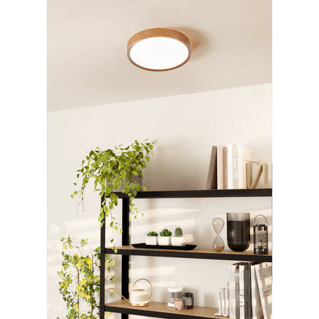Musurita 34cm LED flush ceiling light with natural wood effect rim and white diffuser, shown mounted on a ceiling above a black shelving unit with plants and books.