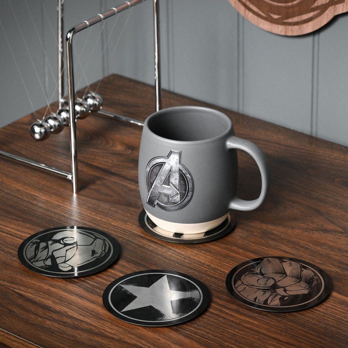 Round metal Avengers coasters in black and silver, three shown on a wooden desk with star, fist, and helmet designs beside a grey Avengers mug on the fourth.