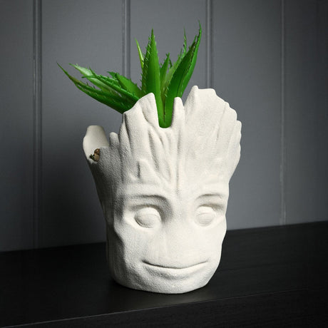 Smiling Groot head ceramic planter in matt cement grey with sculpted bark-textured rim and faux spiky green plant, shown on a dark shelf against a grey panelled wall.