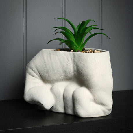 Hulk Fist textured ceramic planter in sandy grey, shaped as a clenched fist with rounded knuckles, holding a faux aloe plant, set against a dark grey panelled wall.