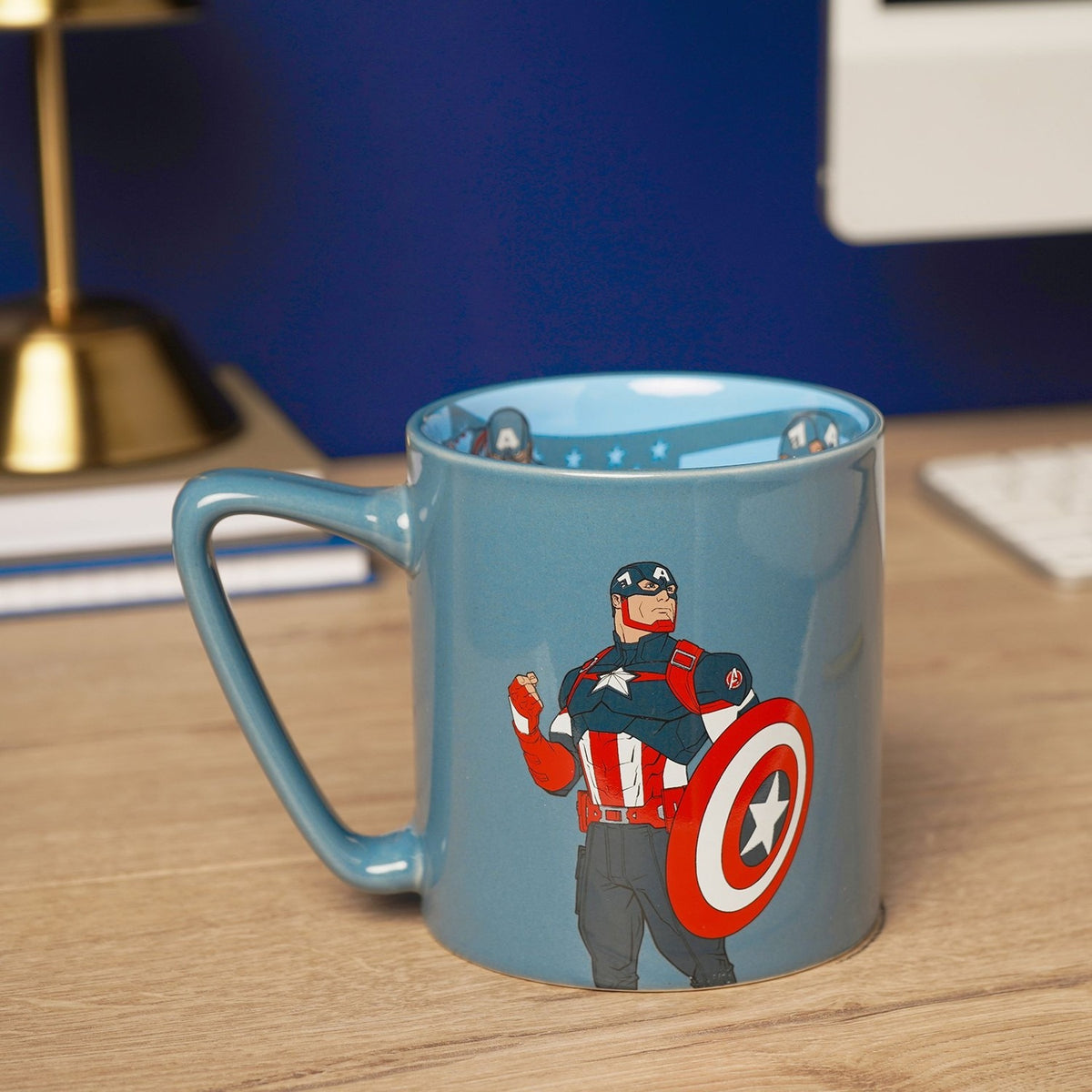 Blue porcelain mug with slate blue glaze featuring Captain America raising a fist beside his shield, light blue interior with printed decal, shown on a desk.