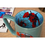Green porcelain Deadpool mug with pale blue inside decal of the character leaping over a cityscape, handle visible, set on a wooden surface.