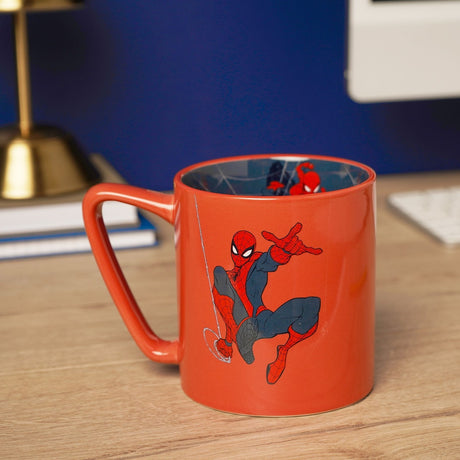 Red porcelain mug with Spider-Man mid-swing printed on the side and a navy city-scene decal inside, shown on a desk with a lamp and keyboard.