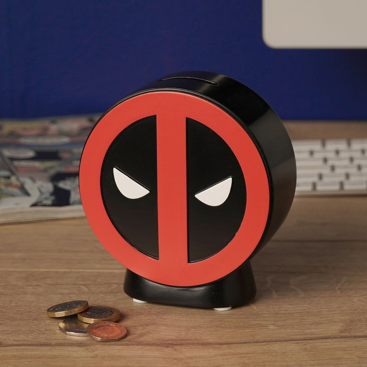 Round Deadpool Emblem resin money box in red and black with moulded relief design, top coin slot, and curved stand, shown on a desk beside loose coins.