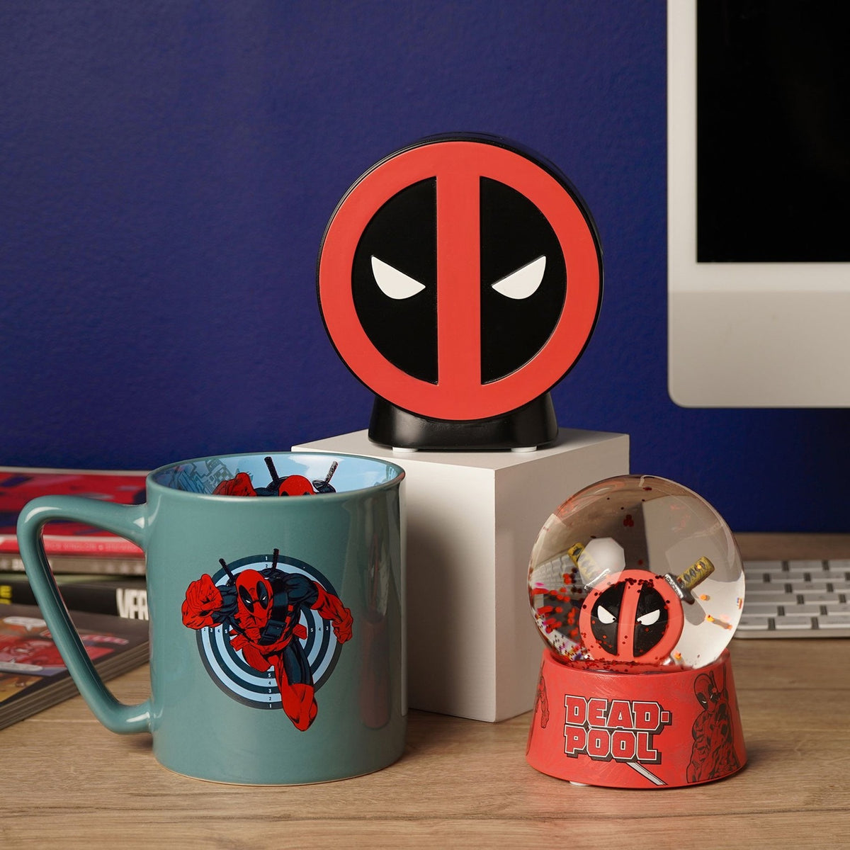 Round resin Deadpool emblem money box in red and black on a stand, shown on a desk with a mug and Deadpool snow globe.
