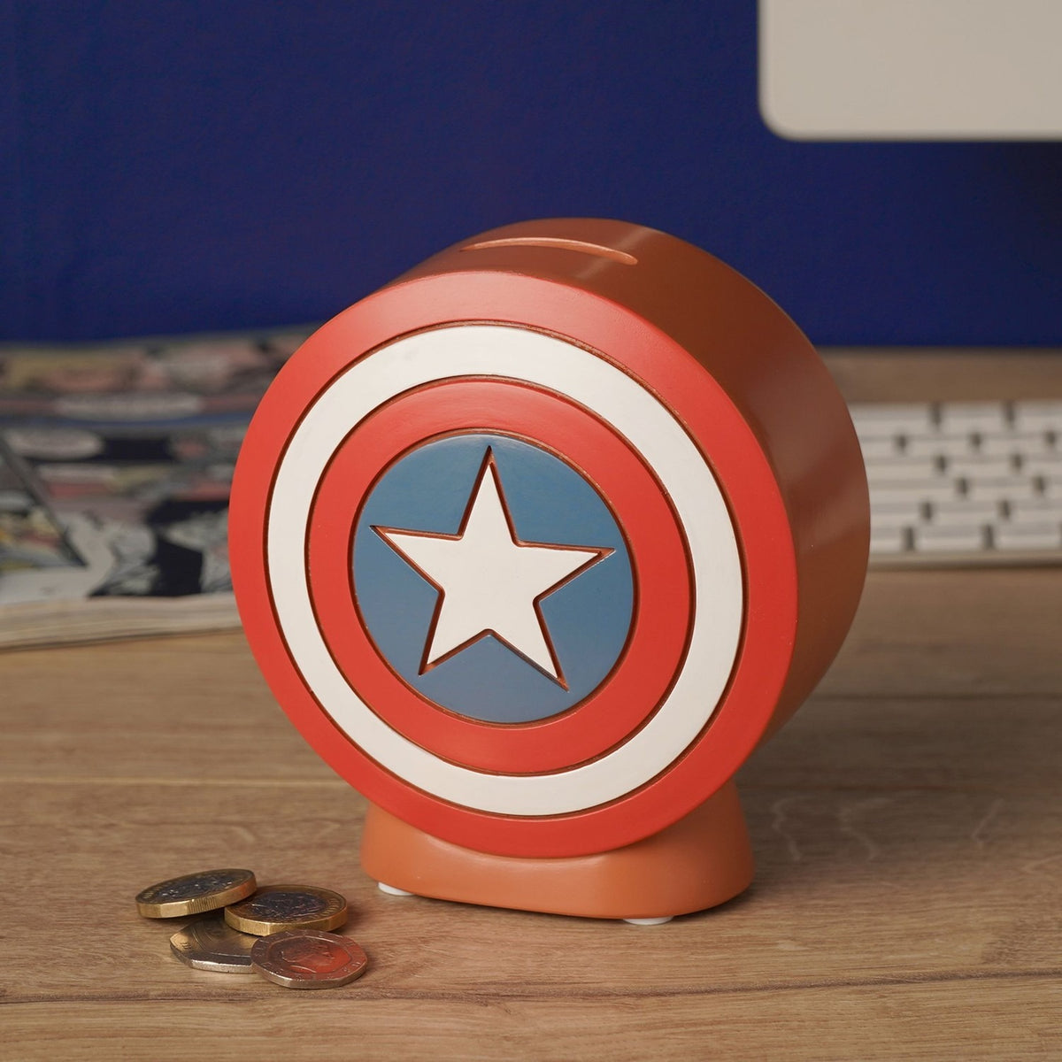 Round Captain America Shield money box in red and white resin with blue centre and white star, displayed on a desk beside loose coins.
