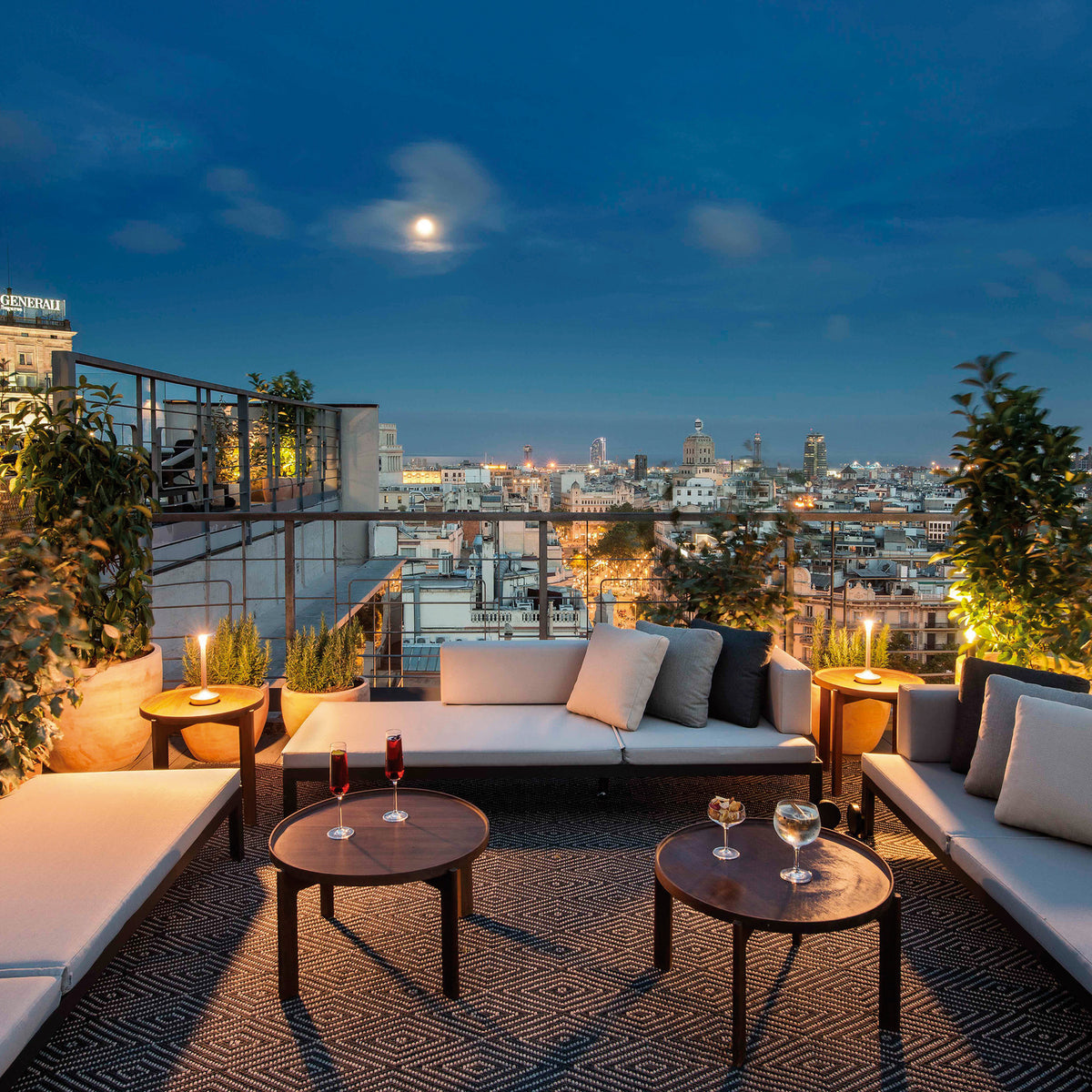 Nak aluminium day and night sun lounger styled as a low sofa with cream cushions on a rooftop terrace overlooking a city skyline at dusk.