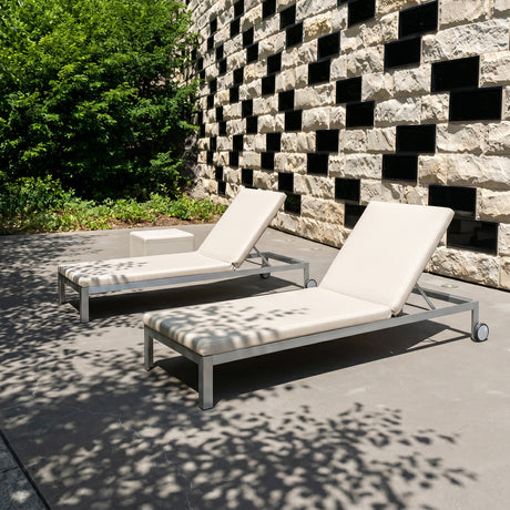 Two Nak aluminium sun loungers with cream cushions and adjustable backrests on a patio, set against a stone wall with greenery.