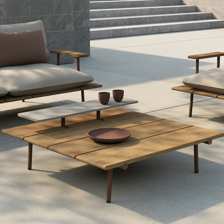 Nami square outdoor coffee table with iroko wood slatted top and raised grey stone second tier on slim aluminium legs, shown on a patio with armchairs.
