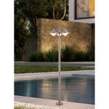 Nasvanne outdoor floor lamp in stainless steel with a tall slim column and three curved arms, each with a metal hood over an opal glass globe, poolside at dusk.