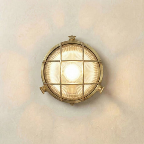 Small Cabin round bulkhead wall light in raw brass with ribbed glass lens, crossed brass cage guard, and thumb-turn lugs, lit against a plain wall.