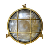 Grand Cabin round bulkhead light in raw brass with grid cage over ribbed glass, three mounting lugs, IP64 outdoor-rated, shown on a plain white background.