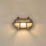 Palaemon Eyelid Bulkhead Light in raw brass, oval shape with ribbed glass lens, guard bars, and brass hood, illuminated on a plain light wall.