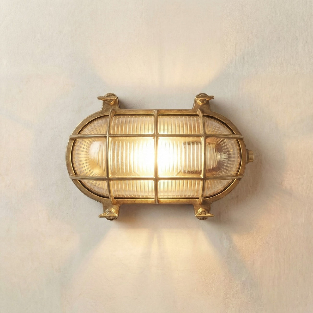 Horatio Oval Bulkhead Light in raw brass, IP64 outdoor wall light with ribbed glass diffuser and crossed brass cage bars, lit against a plain light background.