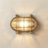 Horatio Oval Bulkhead Light in raw brass, IP64 outdoor wall light with ribbed glass diffuser and crossed brass cage bars, lit against a plain light background.
