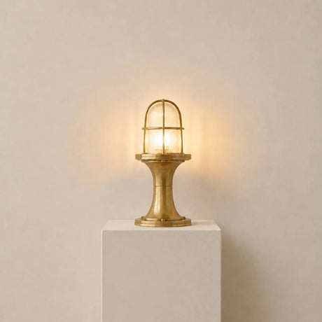 Wellington Outdoor Post Light in raw brass, nautical-style with ribbed glass dome, brass guard, and flared pedestal base, lit and displayed on a plinth against a plain light background.