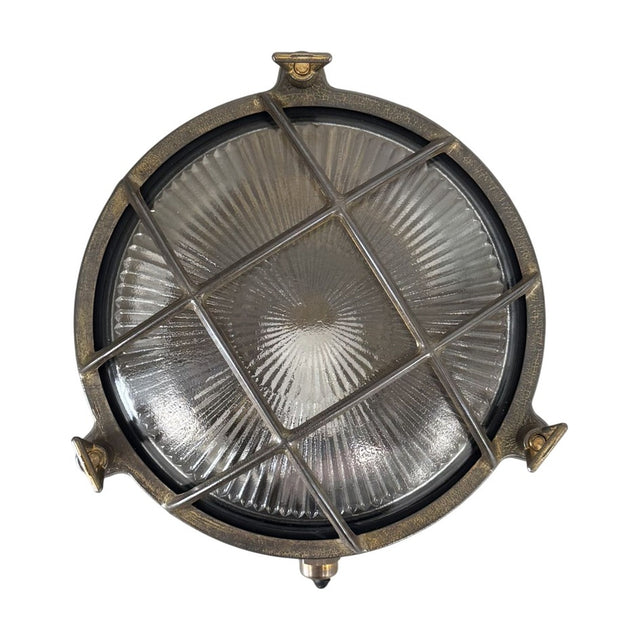 Small round bulkhead light in antique brass with cross grille over ribbed glass, nautical porthole style, shown on a plain white background.