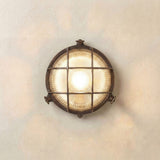 Small Cabin Round Bulkhead Light in antique brass, IP64, with cross grille over ribbed glass, lit, mounted on a plain neutral wall.