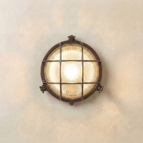 Small Cabin Round Bulkhead Light in antique brass, IP64, with cross grille over ribbed glass, lit, mounted on a plain neutral wall.