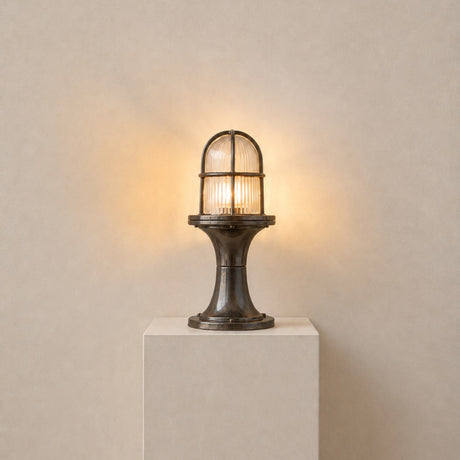 Wellington Outdoor Post Light IP54 in antique brass, with a ribbed glass dome inside a brass cage on a waisted pedestal base, lit, on a plinth against a plain neutral background.