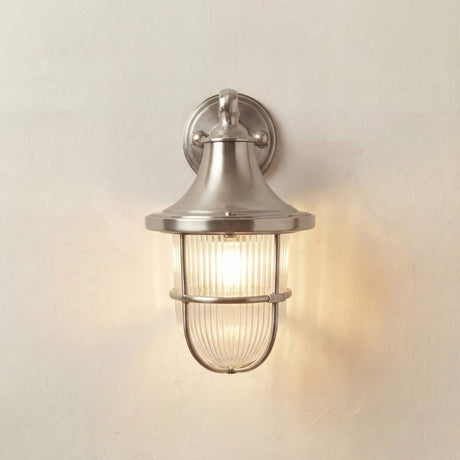 Tiller Outdoor Wall Lantern in satin nickel, featuring a flared hood, ribbed glass, and protective cage on a wall-mounted arm bracket, shown lit against a plain background.