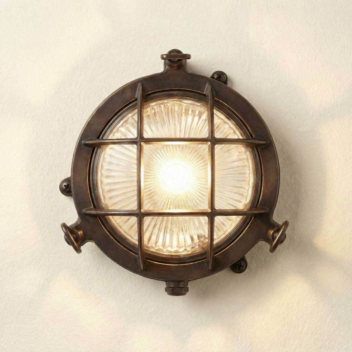 Mini Compass Guard round bulkhead light in antique brass, with crossed-bar grid guard over domed ribbed glass and rim thumb screws, lit against a plain light background.