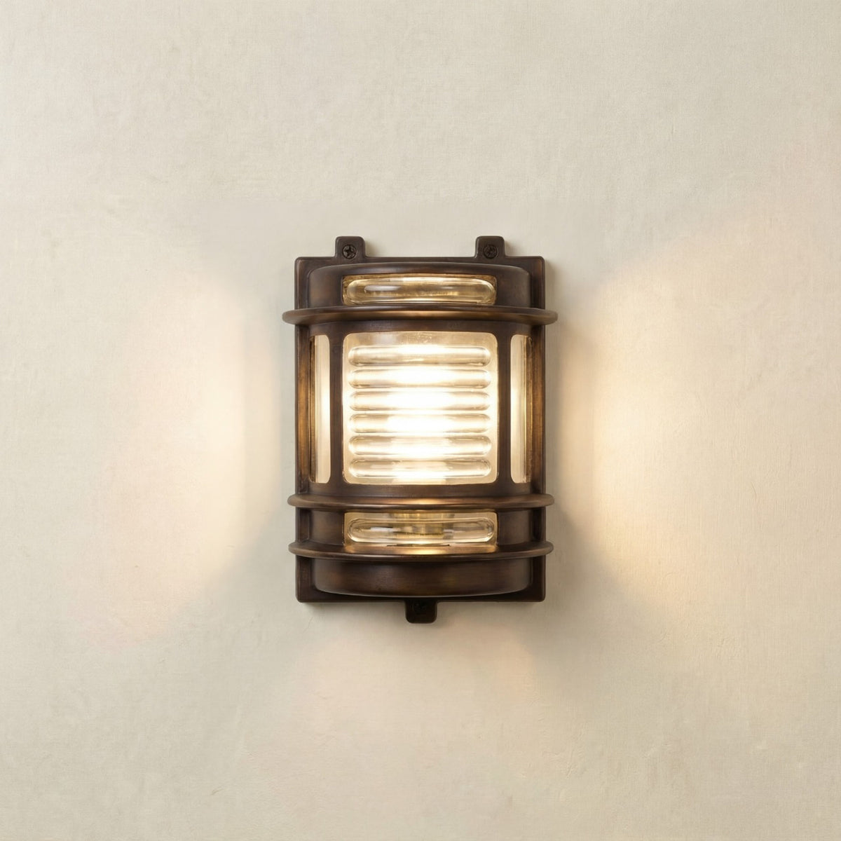 Cargo Outdoor Wall Light in antique brass with ribbed glass panel, lit on a plain light wall, IP64-rated bulkhead fixture with brass bands and side windows.