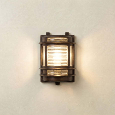 Cargo Outdoor Wall Light in antique brass with ribbed glass panel, lit on a plain light wall, IP64-rated bulkhead fixture with brass bands and side windows.