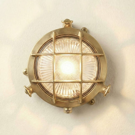 Mini Compass Guard Round Bulkhead Light in raw brass, IP44 rated, with a crossbar grid guard over ribbed glass lens, illuminated, on a plain light background.