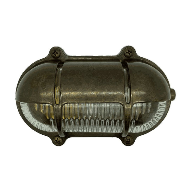 Palaemon North oval eyelid bulkhead light in antique brass, with curved hood shading ribbed glass, brass guard bars and corner fixing lugs, shown on a plain white background.