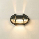 Palaemon North oval eyelid bulkhead light in antique brass, with ribbed glass, brass guard bars, and corner fixing lugs, illuminated on a plain wall background.