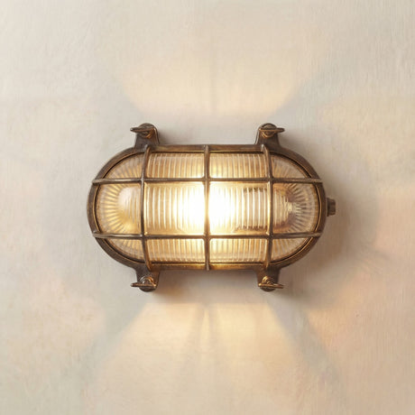 Horatio oval bulkhead light in antique brass, IP64 rated, with ribbed glass diffuser and brass cage lit on a plain wall background.