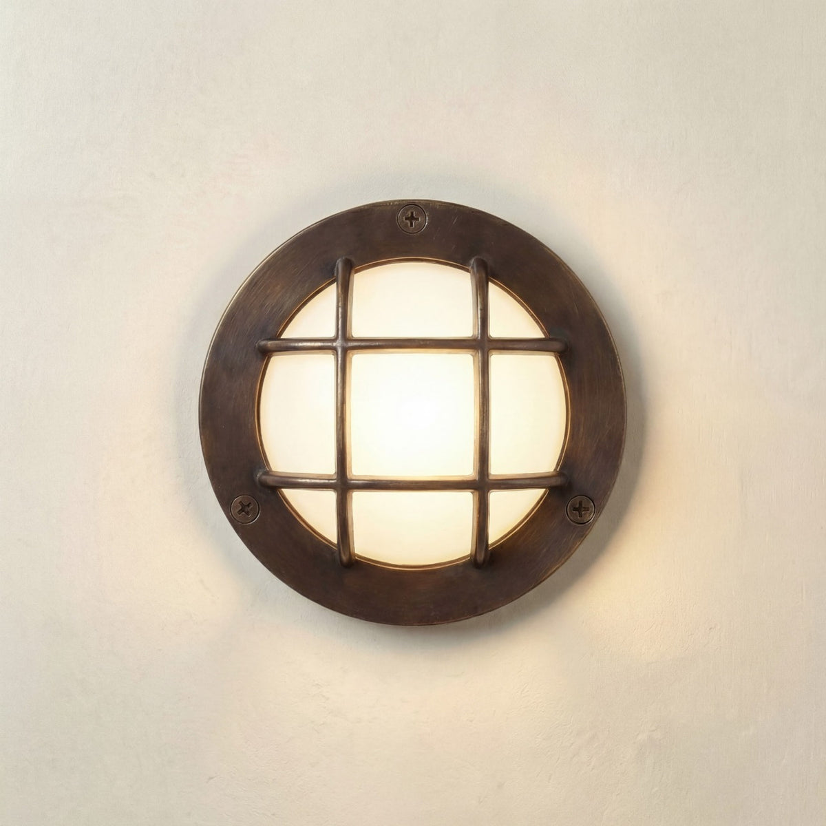 Marina Guard Circular Bulkhead Light in antique brass, round wall fixture with a cross-bar grid guard over a frosted glass diffuser, mounted on a plain light wall.