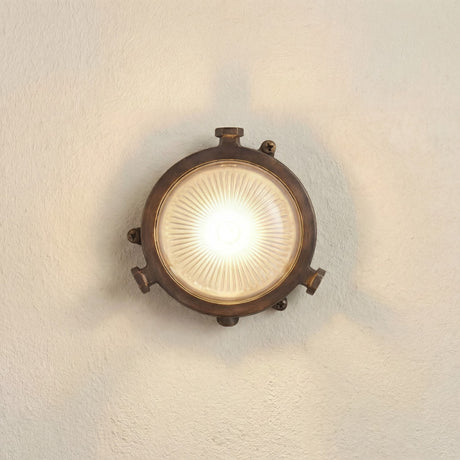 Mini Compass round bulkhead light in antique brass, lit on a wall, with ribbed frosted glass, porthole-style rim and visible mounting lugs, set against a plain light background.