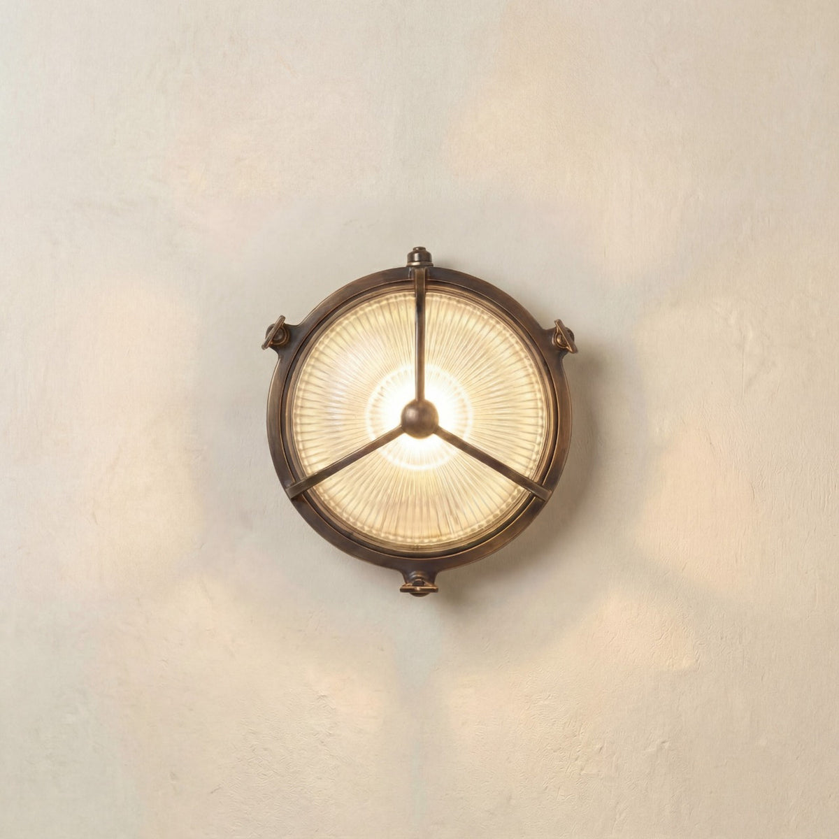 Trident Round Bulkhead Light in antique brass finish, with a three-arm guard over ribbed glass lit from within, mounted on a plain light wall.
